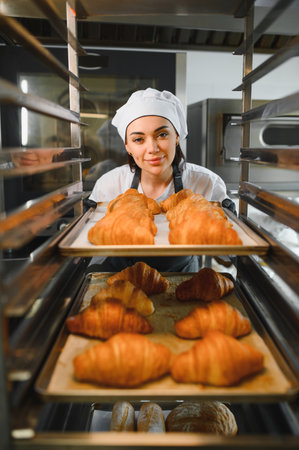 Female baker placing a tray of golden brown croissants into a professional oven, ensuring perfect baking in a bakery or pastry shopの写真素材