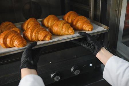 Baker wearing black gloves taking a tray of freshly baked golden croissants out of a professional...