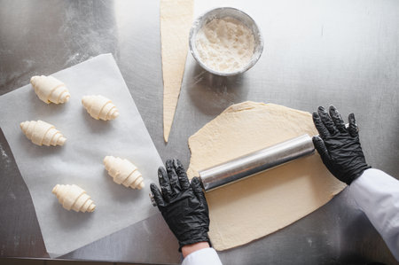 Baker expertly rolling out dough on a metal table, preparing to craft fresh croissants with precise...