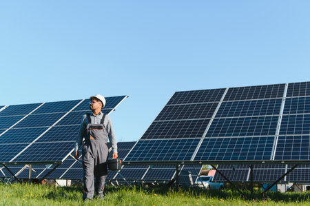 Middle eastern engineer walking among rows of solar panels, carrying a toolbox, overseeing and...