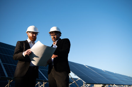 Two engineers wearing hardhats are reviewing blueprints at a solar power plant, showing renewable...