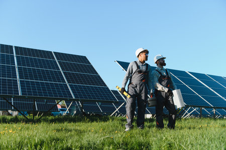 Two engineers holding tools and installing solar panels in a green field with a clear blue sky,...