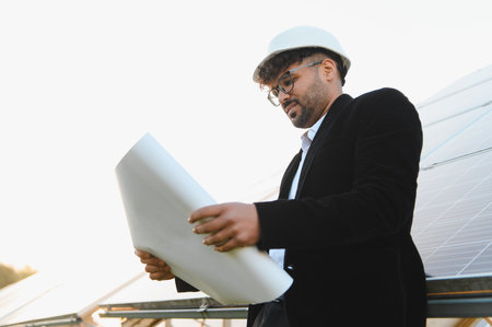 Arab engineer wearing hardhat reviewing blueprints at a solar power plant, showing sustainable energy developmentの写真素材