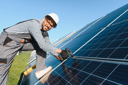 Smiling engineer with safety helmet installing a solar panel using a screwdriver on a sunny day,...