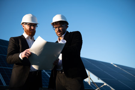 Two engineers wearing hard hats are examining a blueprint in a solar panel farm, discussing...