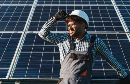 Smiling engineer adjusting hardhat while installing photovoltaic panels at a solar power plant,...