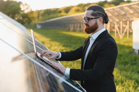 Businessman using laptop analyzing photovoltaic solar panels during sunset in power station,...