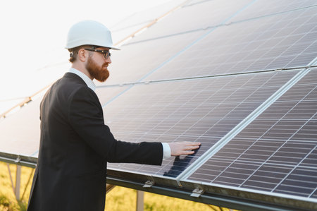 Investor wearing suit and hardhat inspecting a solar panel in a solar farm, touching photovoltaic...