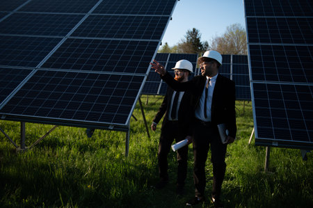 Two engineers in hardhats and suits are walking through a solar panel farm, discussing plans and...
