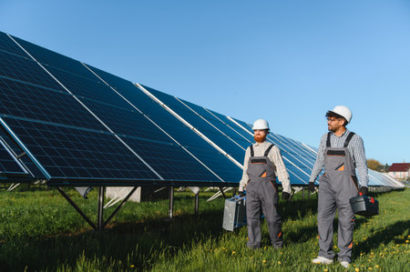 Two engineers carrying toolboxes are walking in a green field near solar panels, contributing to...