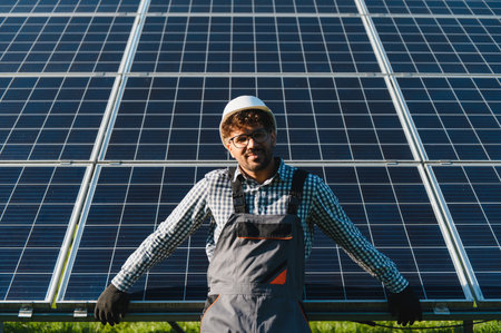 Smiling engineer wearing a hardhat and gloves, installing solar panels at a power plant while...