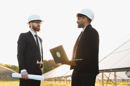 Two engineers wearing suits and hardhats are discussing a project using a laptop in a solar panel farm at sunsetの写真素材