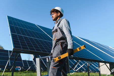 Middle eastern engineer carrying a level tool and walking near solar panels in a large solar farm