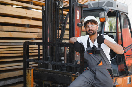 Warehouse worker wearing work overalls and hardhat giving thumbs up while leaning on a forklift near...