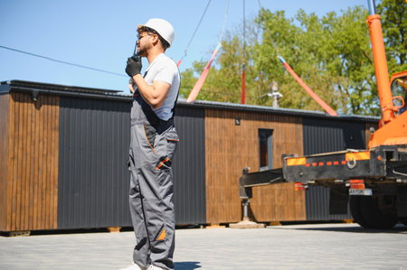 Construction worker using walkie talkie, coordinating modular house transportation onto a truck