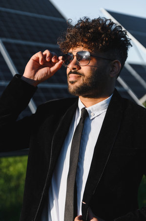 Arab businessman adjusting sunglasses while posing confidently in a solar panel farm, showcasing...