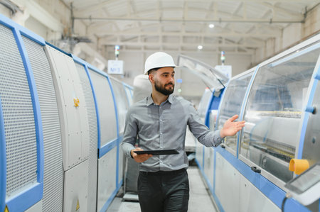 Industrial engineer with white helmet and tablet walking and controlling industrial machinery inside...