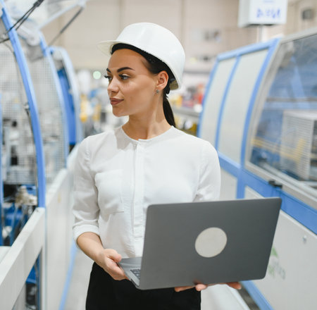 Confident female engineer with white hardhat holding laptop, controlling automated production line...