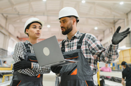 Two factory workers wearing hardhats and overalls are using a laptop and discussing work in a large...