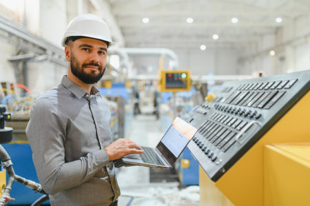 Industrial engineer wearing hardhat using laptop next to control panel while supervising production...