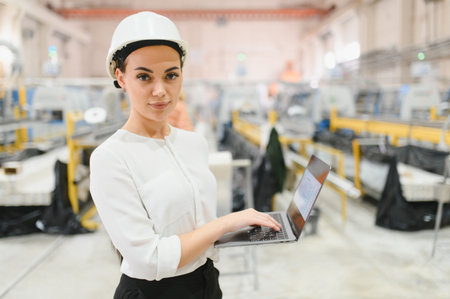 Female engineer wearing a hardhat and using a laptop while overseeing operations in an aluminum and...