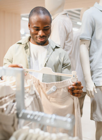 Smiling african american man holding beige trousers on hanger while shopping for new clothes in mallの写真素材