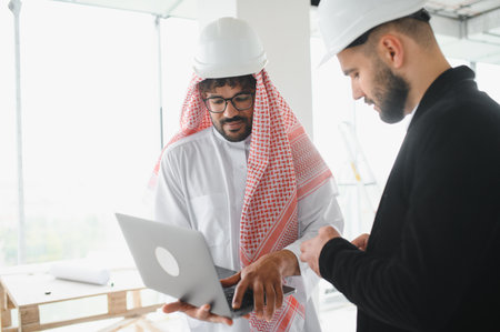 Arab businessman and investor wearing keffiyeh and hardhat using laptop with architect discussing...