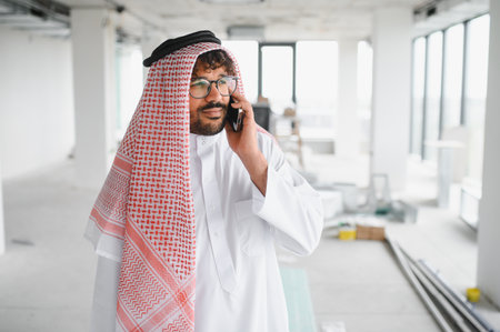 Arab businessman wearing traditional clothing talking on a smartphone at the construction site of his new officeの写真素材