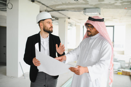 Architect wearing safety helmet showing blueprint to arab investor inside a building under...