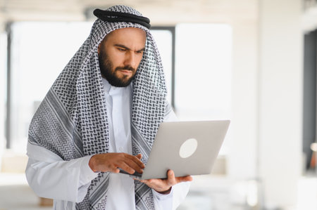 Focused arab businessman wearing traditional clothing using laptop, managing business in bright office spaceの写真素材