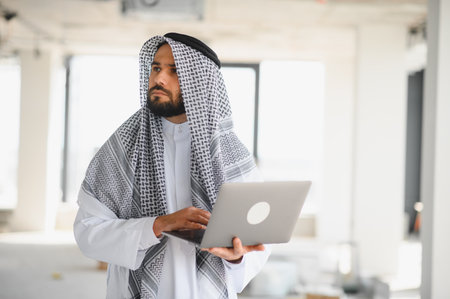 Arab businessman wearing traditional clothing using laptop while inspecting construction site of new business centerの写真素材
