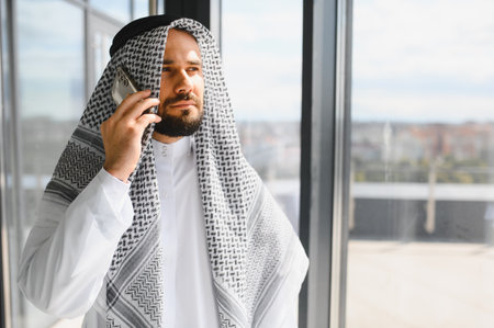 Successful businessman wearing traditional kandura talking on a smartphone in a modern office buildingの写真素材
