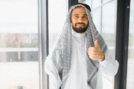 Successful entrepreneur wearing traditional kandura expressing positivity and approval in a bright office environmentの写真素材
