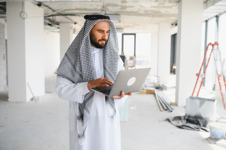 Arab businessman wearing traditional clothing using laptop at construction site of new business center, investments managing and overseeing project developmentの写真素材