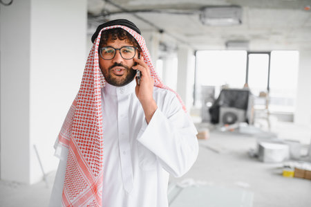 Arab businessman wearing traditional clothing talking on a smartphone while overseeing a...