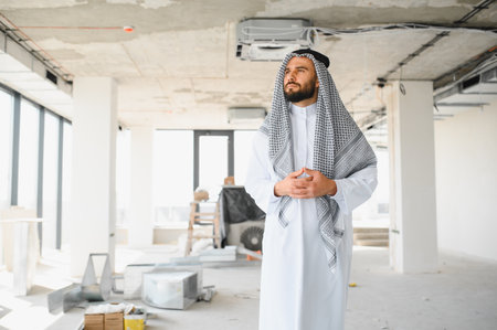 Confident arab businessman in traditional clothing evaluating progress at the construction site of a modern business center, representing investment and developmentの写真素材