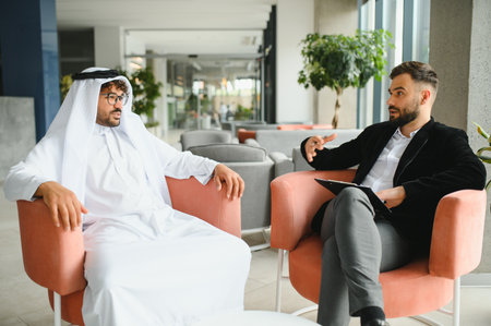 International operations manager discussing business with Saudi partner wearing traditional kandura, showcasing cross cultural communication in a modern office environmentの写真素材