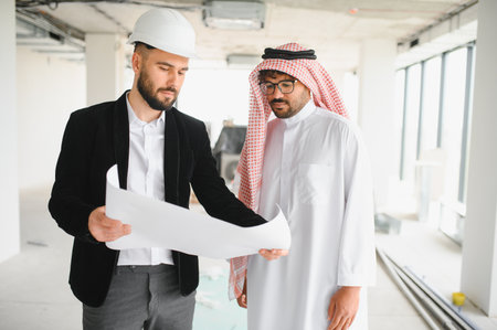 Architect wearing safety helmet showing blueprint to arab investor inside a building under...
