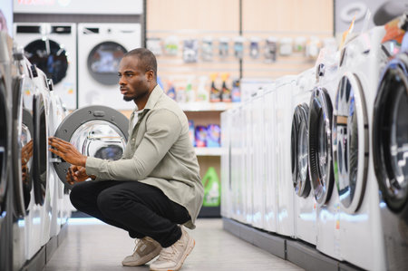 African american man examining washing machines in a shopping mall's electronics store, focusing on choosing the right applianceの写真素材