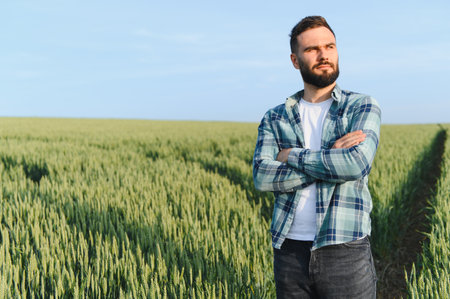 Confident farmer with arms crossed, standing in a lush wheat field, thoughtfully planning the upcoming harvest under a bright blue skyの写真素材