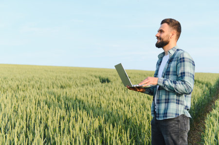 Agronomist using laptop while inspecting wheat field before harvest, ensuring healthy crop growth...