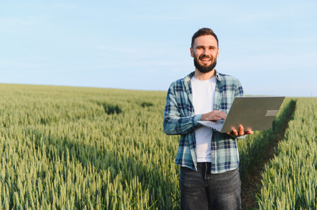 Happy agronomist working on a laptop while inspecting a lush wheat field, preparing for the upcoming...