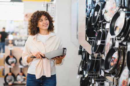 Smiling woman selecting a frying pan while shopping in a vibrant home appliances store, enjoying the process of making a thoughtful purchaseの写真素材