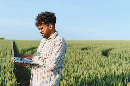 Agronomist is using a laptop in a wheat field, implementing smart farming techniques for efficient...