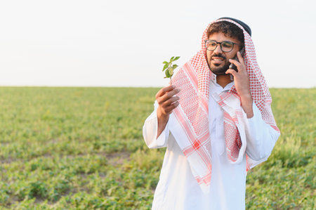 Arab farmer inspects a soybean plant while talking on his phone, showcasing modern agriculture and...