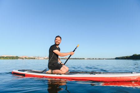 Bearded man enjoying paddleboarding on a beautiful summer day, kneeling on a sup board and paddling...