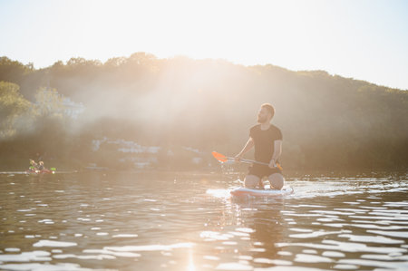 Young sportsman is paddling while kneeling on a sup board on a lake at sunset, enjoying the tranquility of natureの写真素材