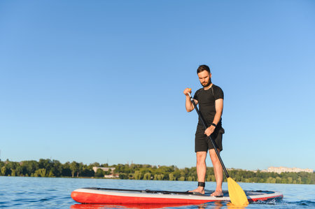 Young sportsman paddling on a stand up paddleboard across a serene lake, embracing the joys of...
