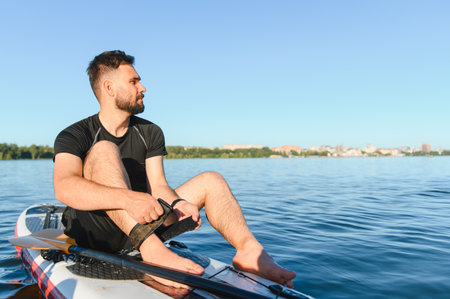 Young sportsman sitting on his sup board, preparing the leash before starting paddling on the lake