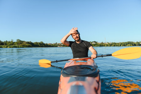 Tired athlete wiping sweat from his forehead while paddling a kayak on a sunny summer day, enjoying the vibrant lake sceneryの写真素材